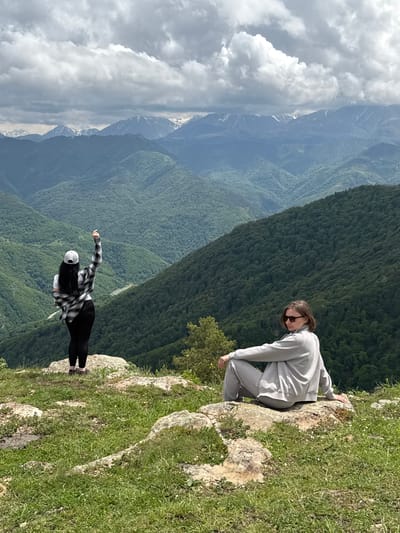Two individuals are situated on a rocky, grassy overlook with a mountainous backdrop. On the left, a person stands with their back to the camera, wearing a white baseball cap, a white t-shirt, a black and white plaid shirt tied around their waist, and black leggings. Their right arm is raised, pointing upwards. On the right, a second person is seated on a rock, facing left, wearing a light gray tracksuit and sunglasses. They are looking away from the camera. The landscape behind them consists of numerous green, tree-covered mountain ranges that recede into the distance, with snow-capped peaks visible on the horizon under a cloudy sky. The scene appears to be in a natural, elevated location, possibly a viewpoint or hiking spot, consistent with the mountainous terrain of Нижняя Теберда, Russia.