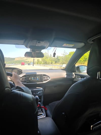 The image shows the interior of a car with a driver's hand on the steering wheel. The view is from the passenger side, looking forward through the windshield. Outside the car, a highway is visible with other vehicles, including a red van and a blue truck. The landscape beyond the highway consists of rolling hills covered in dry vegetation and scattered trees, under a clear blue sky. The car's dashboard features a digital display showing the temperature and climate control settings. The interior of the car is modern, with black leather seats and a black dashboard. A small, dark brown car freshener hangs from the rearview mirror. The photo captures a moment of travel on a sunny day. The provided context suggests this scene is located in the vicinity of Staro Selo, Bulgaria.