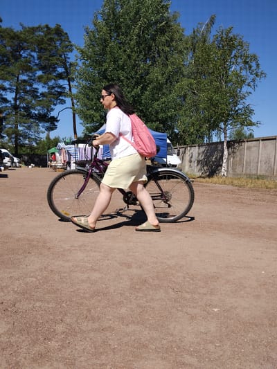 The image depicts a woman walking a bicycle on a dirt ground with a few trees and stalls in the background. She is wearing a white t-shirt, a light yellow skirt, and sandals, with a pink backpack on her shoulders. The bicycle is purple with a black seat and wheels. In the background, there are several stalls with striped awnings, suggesting a market or fair. The sky is clear and blue. The scene appears to be an outdoor event in the city of Irshansk, Ukraine.
