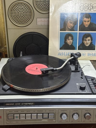 A turntable is playing a vinyl record with a red label on top of a vintage audio system. The label on the vinyl reads "Мелодия" (Melodiya) in Russian. The turntable is a "Unitra" model, specifically the "BE2A-109 STEREO." Next to the turntable is a set of controls including knobs for ГРОМКОСТЬ (volume), БАЛАНС (balance), and ТЕМБР НЧ (bass tone). There are push buttons for ФСНЧ, ОСЛАБ, ФСВЧ, MONO, and other functions. A record cover with the band "Секрет" (Secret) is resting beside the turntable. A speaker cabinet with perforated circular grills and a power indicator is also present. The location Nikolo-Beryozovka, Russia is not discernible from the image.