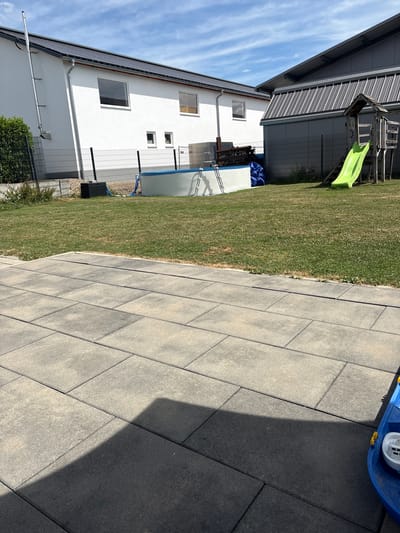 The image shows a backyard in Schwendi, Germany, with a white house and a grey industrial-style building. In the foreground, a paved patio area is visible, with large, rectangular grey pavers. Beyond the patio is a grassy lawn. A round, above-ground swimming pool with a blue liner is situated on the lawn, along with a metal ladder and a blue tarp. To the right of the pool is a wooden play structure with a bright green slide. A dark grey fence encloses the yard, separating it from the neighboring properties and buildings. The sky is blue with scattered white clouds. There are no people visible in the image.