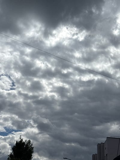 The image shows a cloudy sky above buildings and trees in Olaine, Latvia. A thin dark wire stretches diagonally across the frame from the upper left to the middle right. The clouds are varied in density, with some areas appearing bright and illuminated by the sun, while others are darker and more ominous. In the lower left, the dark silhouette of a tree is visible. Towards the lower right, the tops of several modern buildings are seen, with one structure featuring a railing on its roof. A streetlamp is visible in the lower portion of the frame, and a subtle hint of blue sky can be seen peeking through the clouds in the middle left. The overall impression is of a slightly overcast or approaching storm sky.