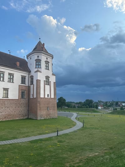The image depicts a view of the Mir Castle Complex in the village of Mir, located within the Zhukhavitski Sel'ski Savet, Belarus. The foreground features a neatly mown green lawn with a paved pathway winding through it. Small yellow flowers are scattered across the grass. The pathway is illuminated by several dark, cylindrical outdoor lamps. To the left, a section of the Mir Castle is visible, characterized by its red brick base and white plastered upper walls. A prominent white tower with a red tiled roof and a weather vane stands prominently on the castle's corner. Numerous windows with dark frames are visible on the castle walls. In the background, a rolling landscape with trees and buildings can be seen under a dramatic sky filled with both bright white clouds and darker, stormier clouds. The overall scene suggests a late afternoon or early evening setting with approaching weather.