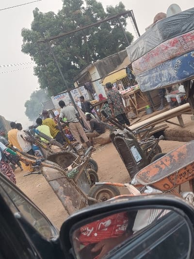 A bustling roadside market scene is captured from a vehicle's perspective. Mattresses are stacked high, while vendors sit or kneel on the ground, displaying fabrics and other wares. Rickshaws line the dusty edge of the road, hinting at transportation options. A rearview mirror provides a glimpse of the car's interior and the driver. Trees and buildings create a backdrop to this vibrant commercial activity.