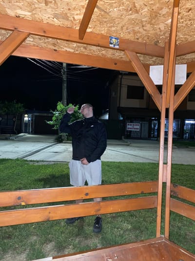 The image depicts a man standing outside at night, partially sheltered by a wooden structure. He is wearing a black jacket and light-colored shorts, with one hand raised to his face. The background shows a street with buildings, a utility pole with wires, and a sign that reads "МАГАЗИН ДРЕХИ И ОБУВКИ" (Shop for clothes and shoes) in Bulgarian. The scene is likely in the city of Yasen, Bulgaria, given the Cyrillic signage and general appearance of the buildings. The man appears to be alone, and there are no other people or direct interactions visible. The environment is a typical urban or semi-urban setting at night, with artificial lighting illuminating the area. The wooden structure suggests a gazebo or an outdoor seating area.
