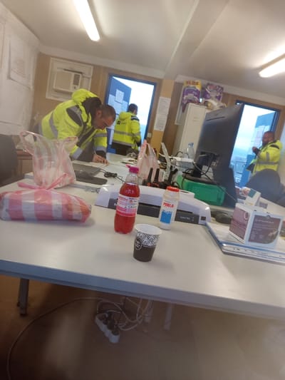 The image displays an indoor scene, identified as an office or break room located in Drugan, Bulgaria. Four adult males are present. All individuals are wearing high-visibility yellow and black work jackets.

In the foreground, a male with short hair and a beard is leaning forward over a light-colored desk, his attention directed towards an object on the desk surface. On this desk, from left to right, are a rectangular item wrapped in red and white striped plastic, a clear crumpled plastic bag, a red plastic bottle containing a red liquid, a white plastic bottle labeled "PERLA" with blue lettering, and a dark-colored disposable coffee cup with a light patterned design. Further along the desk are a black electronic device (possibly a laptop), various black cables, a white office machine (possibly a laminator or scanner), a black computer monitor, a clear plastic water bottle, a stack of blue folders, and a small dark-colored cardboard box.

In the mid-ground, a second male, facing away from the camera, stands near an open blue door, revealing a bright, overcast outdoor environment. A third male is partially visible to the left, also leaning over the same desk. A fourth male is visible in the background on the right, standing near a second open door, looking outward.

The room features a white ceiling with exposed fluorescent tube lighting. The walls are predominantly light-colored, with some sections appearing to be wood-paneled. An air conditioning unit is mounted on the left wall. Papers are affixed to various wall surfaces. A white two-drawer filing cabinet is situated against the back wall, topped with several plastic bags containing items. Below the main desk, a white power strip with multiple outlets and tangled white cables is visible on the floor.