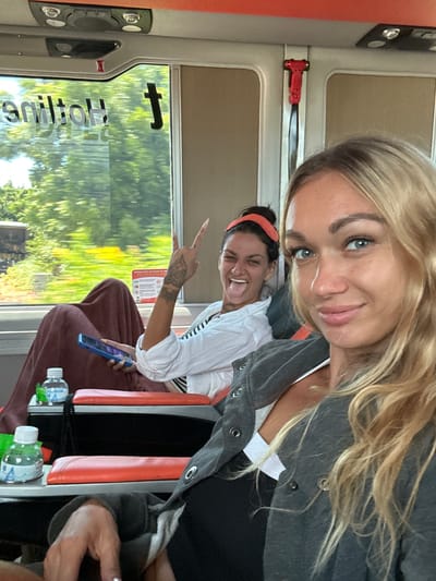 Two women are seated on a bus, traveling through Hòa Thắng, Vietnam. The woman in the foreground, with blonde hair, is looking at the camera and smiling. She is wearing a grey jacket over a white top. The woman behind her has dark hair and is sticking her tongue out at the camera, making a peace sign with her fingers. She is wearing a white shirt and is covered with a maroon blanket. The bus window shows a blurry green landscape with trees. There are bottles of water on a tray between the seats. A red safety hammer is visible on the wall of the bus. The bus appears to be moving.