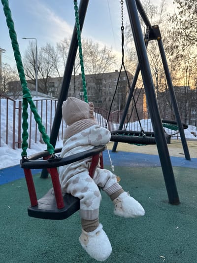 An infant is seated in a red and black toddler swing on an outdoor playground in Sadovaya, Russia. The infant is viewed from behind, facing away from the camera, and occupies the left side of the frame. The infant wears a light brown knitted bonnet with ruffled trim, a light beige one-piece outfit with a repeating pattern of light brown hearts and diamond shapes, brown ribbed leg warmers, and white furry booties adorned with small dark stars. A pacifier on a silver chain hangs from the infant's chest.

The toddler swing features green twisted rope supports and is attached to a dark grey metal swing set. Another black net swing is visible further back on the same swing set structure. The ground beneath the swing set is a multi-colored soft playground surface, primarily blue and green. To the left and in the background, accumulated snow borders a white metal fence. Beyond the fence, a street light pole is visible. The distant background features several grey, multi-story apartment buildings and numerous bare deciduous trees. The sky is a pale blue with some wispy clouds. Low, warm sunlight illuminates a section of a distant building on the right, indicating late afternoon or early evening.