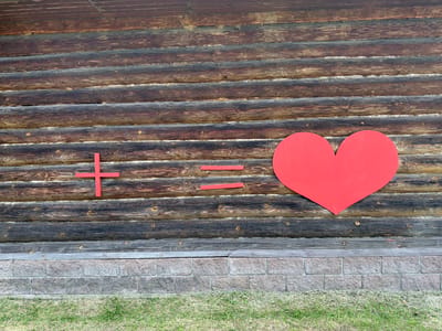 The image displays a section of a weathered log cabin wall in Novy Gorodok, Russia. The logs are horizontally stacked and have a dark, aged appearance with visible wood grain and knot details. Stretched across the logs are three red, rectangular shapes and a large, solid red heart. The shapes are arranged as follows: a plus sign (+) on the left, an equals sign (=) in the middle, and a heart on the right. The plus and equals signs are positioned on the same horizontal log. The heart is slightly lower, resting on the log below the equals sign. Below the log wall, there is a foundation constructed of light gray, rectangular stone blocks. The bottom of the image shows a strip of green grass. The scene is outdoors and appears to be daytime with even lighting. There are no people visible in the image.