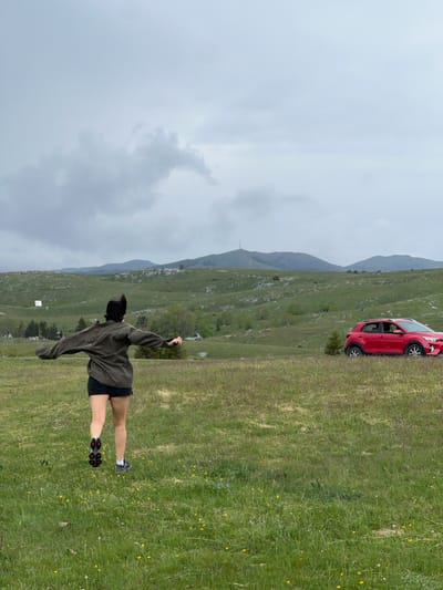 The image depicts an outdoor scene in what appears to be the rural area of Pišče, Montenegro. A young person, viewed from the back, is standing on a grassy slope with outstretched arms. The individual is wearing a dark-colored hoodie, black shorts, and athletic shoes with white socks. A red hatchback car is parked further up the slope. Behind the person and the car, a landscape of mountains and hills stretches into the distance under an overcast sky. The immediate foreground is covered with green grass and wildflowers. The lighting is soft, indicating an overcast day.