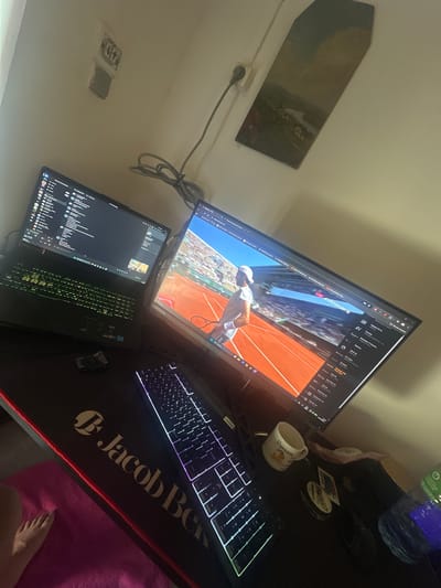 A desk setup is visible in the image. A large monitor displays a tennis match, presumably from the French Open based on the red clay court. Below the monitor is a laptop. To the right of the laptop is a keyboard, sitting on a black mousepad with a logo. A coffee mug is on the desk, near the keyboard. Also visible on the desk are a water bottle and other small objects. Part of a person's foot is in the lower right corner of the image. The location is indicated to be Savigny-en-Véron, France. A framed picture is hanging on the wall. An electrical outlet is on the wall as well.