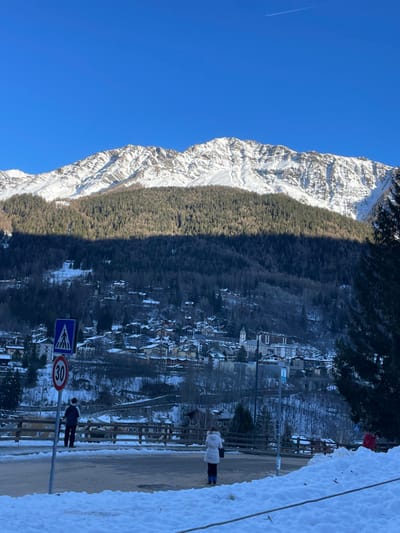 The image captures a panoramic view of a snow-covered alpine landscape, likely in Courmayeur, Italy. A majestic, rugged mountain, heavily dusted with snow and featuring rocky peaks, dominates the left side of the frame under a clear, bright blue sky. A faint airplane contrail is visible high in the sky.

Midway down the mountain, a dense, dark green forest of evergreen trees forms a prominent horizontal band, separating the sun-drenched upper slopes from the shadowed valley below. This band of trees casts a long, distinct shadow across the right side of the image, indicating either early morning or late afternoon.

In the mid-ground on the right, a charming town or village is nestled in the valley, partially obscured by the shadows. Its buildings, many with traditional pitched roofs, are also dusted with snow, giving the settlement a picturesque winter appearance.

In the immediate foreground, a paved road or street runs horizontally. To the right of the road, a snow-covered bank or path extends towards the edge of the frame. Two individuals are visible in this area. One person, wearing a light-colored puffer jacket and dark trousers, stands on the snow, facing away from the viewer and looking towards the town. Closer to the road, another person in dark clothing stands near a signpost.

The signpost displays two road signs: a blue square sign with a white triangle and a pedestrian symbol, and a white circular sign with a red border indicating a "30" speed limit. A brown guardrail or fence runs alongside the road. The overall scene conveys a serene winter day in a mountainous region, characterized by crisp air and dramatic natural beauty.