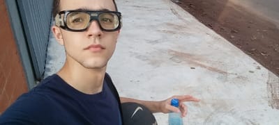 The image is a selfie taken by a young man wearing black safety glasses and a navy blue t-shirt. He is standing on a concrete sidewalk in Encarnación, Paraguay, near a street with a dirt shoulder. He is holding a blue plastic bottle with his right hand, and a black and white object, possibly a skateboard, is visible in the lower right corner. His left hand is pointing towards the right side of the image.  The image is taken from a low angle, with the sidewalk in the background and the subject in the foreground. The sidewalk is cracked and stained, while the street is covered with dirt and gravel. The background is blurry. The coordinates of the image are latitude -27.325, longitude -55.872.