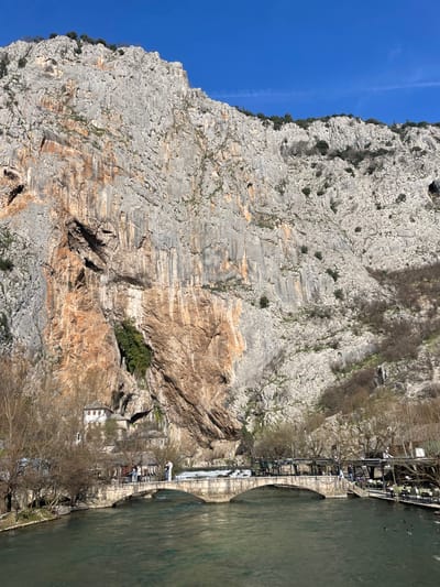 The image displays a panoramic view of the Buna River in Blagaj, Bosnia and Herzegovina, set against a massive limestone cliff under a clear blue sky. In the lower portion, a dark green river flows. A light grey stone arch bridge, featuring multiple arches, spans the river. Several individuals are visible on the bridge; one person stands prominently on the far left side, facing the cliff. On the right bank, an outdoor restaurant area with dark tables, chairs, and overhead coverings is occupied by multiple people. On the left bank, leafless deciduous trees are present. Directly at the base of the cliff and immediately behind the bridge on the left, the Blagaj Tekke (Dervish Monastery) complex is visible, consisting of white-walled buildings with grey roofs, some appearing integrated into the rock face, along with a prominent minaret-like structure. A small waterfall descends from the cliff above the Tekke. The towering limestone cliff dominates the upper two-thirds of the scene, exhibiting textured surfaces with shades of grey, white, and prominent reddish-brown and orange streaks. Multiple dark cave openings are discernible within the cliff face, particularly above the Tekke and to the right. Sparse green foliage and shrubs cling to the cliff's ledges and upper sections. The sky above is uniformly clear and blue, with a faint contrail visible on the far right. Several dark waterfowl are present in the river on the right side.
