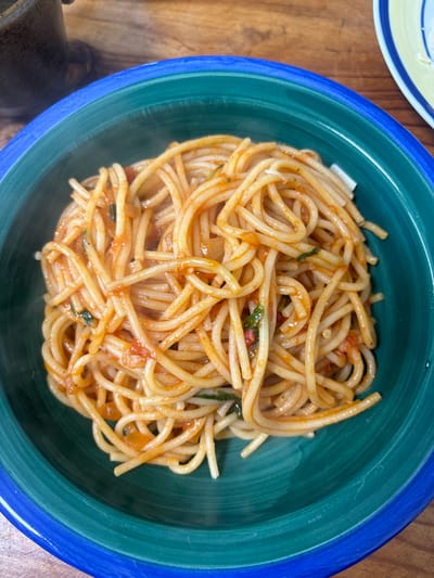A high-angle close-up depicts a steaming bowl of spaghetti coated in a reddish-orange sauce with visible pieces of red tomato, dark green herbs, and lighter-colored ingredients, possibly onion or cheese. The pasta is contained within a deep teal ceramic bowl featuring a distinct blue rim. The bowl rests on a wooden surface exhibiting a grain pattern. A dark brown, possibly ceramic, mug is partially visible in the upper-left background, while a segment of a light-colored plate with yellow and blue stripes is visible in the upper-right background. The scene is located in the city of Menashe Regional Council, Israel.