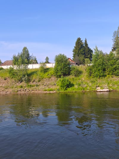 The image shows a wide, calm river flowing in front of a green, grassy embankment under a clear blue sky. The embankment is lined with trees and vegetation, with a white fence and a few houses visible in the background. The water reflects the surrounding greenery and the sky. The scene depicts a peaceful, natural environment, possibly on the outskirts of a rural settlement like Galanino, Russia. There are no people or animals visible in the image. The lighting suggests it is a sunny day, likely during daylight hours.