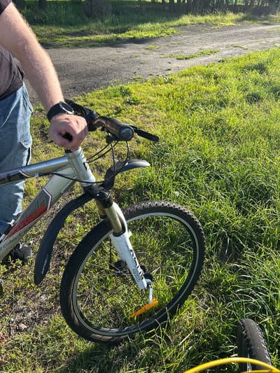 A person's arm and hand are visible in the upper left quadrant of the image, resting on the handlebars of a silver mountain bike. The person is wearing a black t-shirt and blue jeans. The bike is parked on a grassy area next to a dirt path, which is partially obscured by the bike. In the background, trees and more grass are visible. The overall scene suggests an outdoor activity, possibly a break during a bike ride. The location is the city of Bałdowo, Poland.