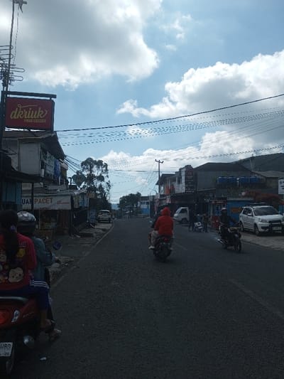 A street scene in the city of Naung Bo Oe, Myanmar, features a paved asphalt road beneath a sky with scattered white clouds. In the foreground on the left, a red scooter carries two occupants: a driver wearing a helmet and a light-colored jacket, and a passenger in a red shirt patterned with characters. Utility poles with numerous overhead wires are prominent along the left and extending across the upper frame. A building on the left displays a red sign reading "dkriuk FRIED CHICKEN" and a partially visible sign below it indicating "UAH PACET". Further down the road, a person in a red hooded jacket rides a dark scooter centrally. On the right side of the road, another individual in dark clothing and a helmet operates a dark scooter. A white SUV and a white van are parked along the right side of the street, where various buildings with multiple stories are visible, some featuring blue water tanks on their roofs. The road recedes into the distance, bordered by additional structures and distant hills or mountains on the horizon. Several indistinct figures are visible near the buildings on the right.