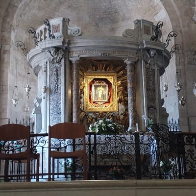 This image depicts the interior of the Cathedral of San Ciriaco (Duomo di Ancona) in Ancona, Italy, specifically focusing on the Altar of the Madonna della Misericordia located in the right transept.

The central subject is an ornate, Baroque-style altar. It features two fluted, dark-veined marble columns (possibly Breccia Sanguigna) supporting a curved architrave. Gold-leafed sunburst rays emanate from a central niche, which houses a framed icon of the Virgin Mary. The icon itself is relatively dark, depicting the Madonna's face and upper torso, adorned with a crown and veil, set within a multi-layered gilded frame. Above the niche, the Latin inscription "REGINA SANCTORUM OMNIUM" (Queen of All Saints) is visible.

Below the icon, on the altar surface, there is an arrangement of white flowers, likely lilies, and green foliage. A single white candlestick with a small, unidentifiable object or a very short candle is present on the right side of the flower arrangement.

In the foreground, a dark, intricately scrolled wrought-iron railing forms a barrier. Two light-brown wooden chairs with simple, unpadded designs are positioned in front of this railing, facing the altar. The floor in the immediate foreground appears to be a light-colored, smooth surface.

The environment indicates a large stone structure. The ceiling above the altar is a vaulted, light-colored stone or brick construction. Numerous silver-colored, ornate hanging lamps, shaped like chalices, are suspended by chains from the ceiling, particularly on the left and right sides of the altar structure. The walls are constructed from light-colored, possibly stuccoed, stone. No individuals are present in the frame.