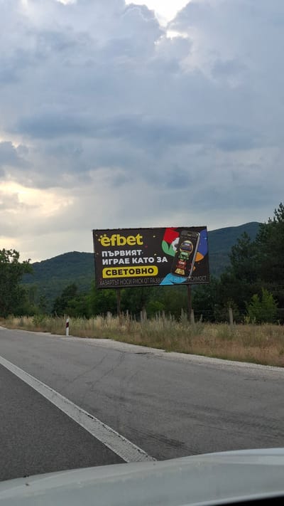 The image is a medium-distance shot of a billboard on the side of a road, likely in Dolno Varshilo, Bulgaria, given the context. The billboard features the "efbet" logo in yellow, followed by the text "пЪРВИЯТ ИГРАЕ КАТО ЗА СВЕТОВНО" (The first plays like for the World) in Bulgarian. Below this, in a yellow highlighted box, it says "СВЕТОВНО" (World). A smaller line of text underneath reads "ХАЗАРТЪТ НОСИ РИСК ОТ РАЗВИВАНЕ НА ЗАВИСИМОСТ" (Gambling carries the risk of developing addiction). The billboard also displays a smartphone image showing a soccer ball and a trophy, with country flags like USA, MEX, and CAN visible. The background shows a hilly, tree-covered landscape under a cloudy sky with patches of sunlight breaking through. The road in the foreground is asphalt with a white line marking. There are no people visible in the image.
