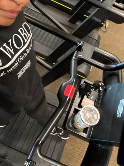 The image depicts a partial overhead view of an individual within a fitness center located in Plainsboro Township, United States. The person, whose lower face and upper torso are visible on the left, is wearing a black long-sleeved shirt with white text, reading "WOBD" and "Law & Order Special Victims Unit." The subject is positioned on or near a black Technogym fitness machine, likely a stepper or elliptical, featuring black handlebars and a red emergency stop button. On the machine's console tray, a pair of black sunglasses, a black car key fob, a small light-colored oval stone, and an insulated cylindrical cup with a clear lid and a colorful aquatic design are visible. Multiple additional black fitness machines with yellow accents are present in the background, situated on a light brown carpeted floor. No specific actions are observable beyond the presence of the individual and objects in the gym environment.