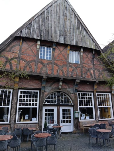An exterior view captures a multi-story, half-timbered building with a steep gable, located in Burgsteinfurt, Germany, under an overcast sky. The upper gable section is clad in vertical, weathered wooden planks. Below this, the second story consists of red brick infill framed by dark wooden beams forming X-patterns and decorative arches. This level features two rectangular windows with white frames and multiple panes. The ground floor maintains the brick and timber construction, presenting three large rectangular windows with white multi-pane frames. The central entrance features a white double door with multiple panes, topped by an arched transom window, also multi-paned. A black wrought-iron sign, partially visible, displaying "SCHWARZ" is mounted above the main entrance. To the right of the door, a small green wall-mounted mailbox or information box is present. A light wooden bench is positioned below the central ground-floor window, which contains a white vase with branches, a large cream-colored bunny figurine, and printed notices. A small potted evergreen plant stands to the right of the door. The forecourt is paved with cobblestones, upon which multiple outdoor tables with brown wooden tops and dark grey woven chairs are arranged. Sparse tree branches with light green leaves are visible on the left and right sides of the frame. No individuals are present in the scene.