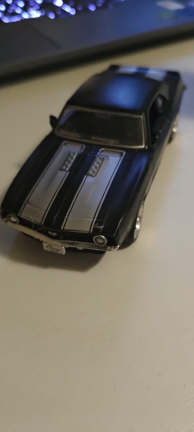 The image shows a close-up, high-angle shot of a black toy car with silver racing stripes on its hood. The car has silver trim around its headlights and grille, and a silver license plate with the word "Camaro" on it. The car is placed on a light-colored, flat surface, possibly a desk or table. In the background, out of focus, is a dark object with colorful lights, suggesting it could be a computer keyboard or similar electronic device. There is no indication of the location being in Skopin, Russia.