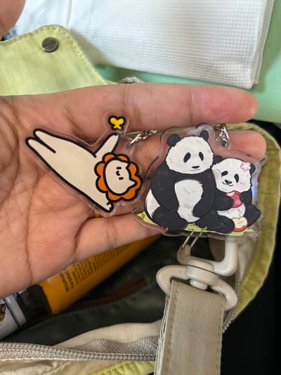 A person's hand is holding two acrylic keychains with metal clasps. The left keychain features a cartoon of a white figure with a round, orange, flower-like head and a yellow butterfly above. The right keychain depicts two pandas sitting on green grass with an apple. The keychains are attached to a light-colored bag with a tan strap. In the background, the bag's fabric is visible, along with a white textured surface and a yellow tube. The scene appears to be an indoor setting, possibly a home or a store.