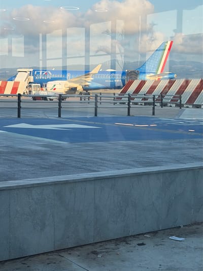 The image shows an airplane parked on a runway. The airplane is blue with white text that reads "ITA AIRWAYS" and an Italian flag on its tail. The runway is marked with blue and white lines. In the background, there is a building with red and white striped panels. The sky is blue with some clouds. The image appears to have been taken during the daytime. The scene is likely at an airport in Lamezia Terme, Italy, as indicated by the location context. There is a reflection of what appears to be a person's eye in the glass in the foreground, suggesting the photo was taken from inside a building looking out.