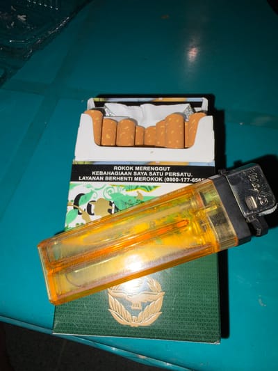 The image shows a close-up, high-angle shot of a pack of cigarettes and a transparent orange lighter placed on a green surface. The cigarettes are visible inside the open pack, with their orange filters facing upwards. The lighter is positioned diagonally across the cigarette pack. The green surface appears to be a table or a similar flat object with a subtle textured pattern. The background is a teal-colored surface, possibly a wall or another table. The lighting in the scene is dim, creating shadows and highlighting the objects. The overall impression is that of a still life composition. The provided information about the location in Tupah, Indonesia is not visually supported by the image content.