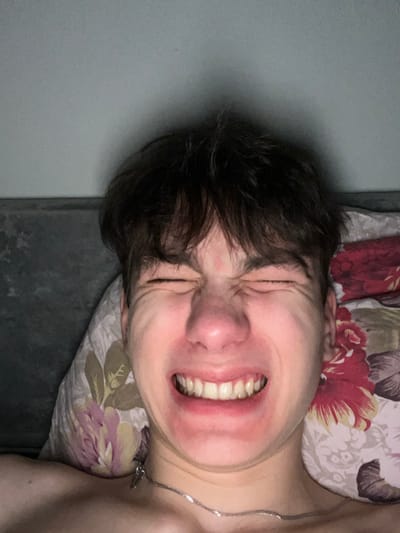 A close-up, low-angle photograph depicts a single young male lying down indoors. The individual, with short, dark, tousled hair, is centered in the frame. His eyes are tightly shut, his nose is crinkled, and his mouth is wide open in a pronounced grimace, baring both upper and lower teeth. His face appears flushed, particularly around the nose and cheeks. A thin silver chain necklace with a small pendant rests on his exposed neck and upper chest. He is positioned against or on floral-patterned pillows: one on the left features purple/pink flowers and green leaves on a light background, and another on the right displays prominent red flowers and green leaves. A dark gray or charcoal-colored surface, possibly a headboard or wall, is visible in the upper background. There are no identifiable features within the image that indicate the specific location within Zainsk, Russia.