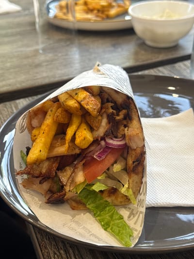 A close-up, high-angle shot captures a generously filled Greek-style wrap, likely a gyro or souvlaki, served on a dark, glossy plate or tray. The wrap, held together by white paper packaging, is packed with grilled meat (appearing to be chicken or pork), golden-brown french fries, shredded lettuce, sliced red onion, and a wedge of red tomato. The pita bread is visibly grilled and slightly charred.

To the upper left, out of focus, a small white bowl, possibly containing a dipping sauce, and a blurry pile of additional french fries are visible. Behind the main subject, a white paper napkin rests on the dark tray. The overall setting is a casual dining environment, suggested by the fast-casual food presentation and simple tableware. The surface beneath is a light-colored wooden table.

The paper packaging for the wrap features black text and graphics. Visible partial words and letters include "TO," "OMA," and phrases like "ILON CV9" and "ME... E," suggesting brand names, slogans, or possibly Greek script on the wrapper, aligning with the style of cuisine. The scene depicts a prepared meal, possibly for consumption in a casual restaurant or as a takeaway item, during what appears to be daytime due to the bright, even lighting. No people are visible, and no specific emotions can be discerned. The location context is Rushmoor, United Kingdom, where such international cuisine is commonly available.