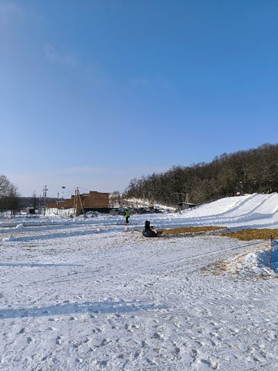The image displays a snow-covered landscape in Prybuzke, Ukraine, under a clear blue sky. In the mid-ground, a snow-covered field shows numerous tracks from foot traffic and sleds. To the left, several brick-colored buildings are visible behind a low white fence, alongside utility poles and power lines. Further right, a densely forested hill rises, with a prepared snow tubing or sledding slope featuring multiple parallel tracks descending towards the foreground. A parking area with vehicles is situated between the buildings and the slope. Two individuals are present: one person, wearing a bright green or yellow jacket, stands upright in the mid-ground, facing away from the viewer. Another person is seated on a dark-colored inflatable snow tube, facing left, with a pink item visible near them. A blue mesh safety net is partially visible on the far right edge of the foreground. The overall scene suggests a winter recreational area.
