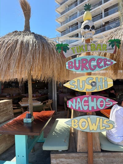 The image captures a daytime scene at a tiki bar in Cala Rajada, Spain, identified by the prominent "TIKI BAR" sign. Various surfboard-shaped signs hang indicating menu items: "BURGERS," "SUSHI," "NACHOS," and "BOWLS". The bar is constructed with thatched roofing and wooden supports.

The scene also depicts outdoor seating, with a table and chairs positioned within the frame. A person is seated at the table, partially visible. The background reveals a glimpse of the building behind the tiki bar.