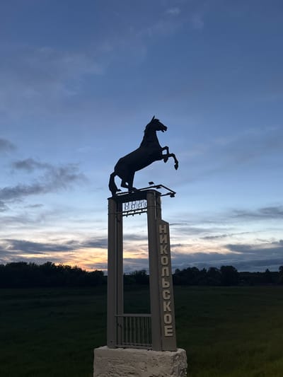 A silhouette of a rearing horse statue stands atop a pillar with the Cyrillic word "НИКОЛЬСКОЕ" vertically inscribed on its side. The statue is set against a twilight sky with streaks of orange and blue from the setting sun. Below the pillar, a dark green, grassy field stretches towards a treeline. The scene appears to be an outdoor monument or landmark.