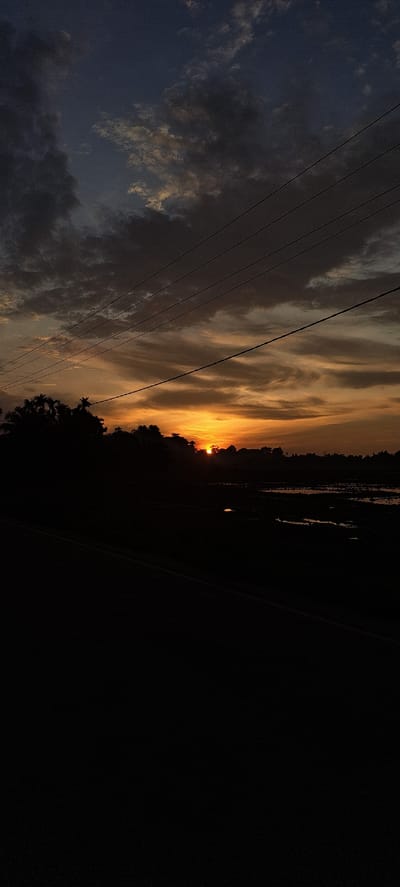 This is a vertically oriented, wide-angle photograph of a sunset in Ngamatangubi, Indonesia. The sun is setting on the right side of the frame, casting a warm, orange glow across a cloudy sky. The clouds are wispy and streaked, with some appearing dark and heavy, while others are illuminated by the setting sun.

In the foreground, several power lines stretch diagonally across the sky from left to right. The land below the horizon is mostly obscured by darkness, but appears to be silhouetted with trees and possibly some structures. A faint light can be seen in the distance over the water, potentially a streetlight or a distant building.

The time of day is sunset, as indicated by the low position of the sun and the warm colors in the sky. The weather appears to be partly cloudy, with a mix of dark and illuminated clouds. The location cues of power lines and silhouetted trees suggest a semi-rural or suburban setting. The overall mood of the image is serene and peaceful, with the dramatic colors of the sunset creating a sense of awe. There is no visible text in the image.