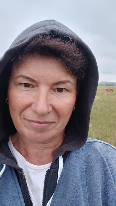 A medium shot shows a woman with brown hair and brown eyes wearing a dark grey hooded sweatshirt over a white t-shirt. She is smiling slightly and looking at the camera. The background is a grassy field with a few blurred horses in the distance. The sky is overcast. The woman is positioned in the foreground, with the field and horses out of focus in the background. The image appears to have been taken outdoors.