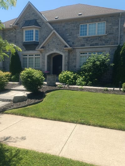 This image shows the front of a large, two-story stone-clad house. The house has a dark-shingled roof, multiple windows with white frames, and a prominent bay window on the upper floor. A covered entryway with an arched opening leads to the front door. The landscaping in front of the house includes a well-maintained lawn, various shrubs and bushes, and a stone pathway. The scene is set in Oakville, Canada, on a sunny day.