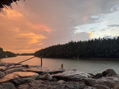 The image depicts a serene river scene at dusk in Rhieng Krueng, Indonesia. The sky is filled with a dramatic display of clouds, with hues of orange, pink, and gray. A dense line of palm trees borders the right side of the river, creating a dark, lush backdrop. The foreground is dominated by large, wet rocks and gravel, suggesting the riverbank. A wooden post with a rope tied to it stands in the water, possibly indicating a marker or part of a fishing setup. The river itself is calm, reflecting the muted colors of the sky, and appears to have a slight ripple effect indicating rain is falling or has recently fallen. The overall atmosphere is tranquil and natural.
