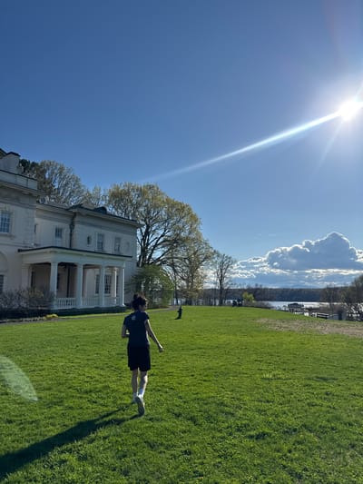 The image depicts a person in a dark-colored shirt and shorts lying on a green lawn. The person is oriented horizontally in the frame. A large white building with columns is partially visible behind them to the left, and a line of trees and a body of water are in the background. The sky is bright blue with a prominent sun flare in the upper left corner and some clouds visible to the right. The location is in the Town of Red Hook, United States of America.