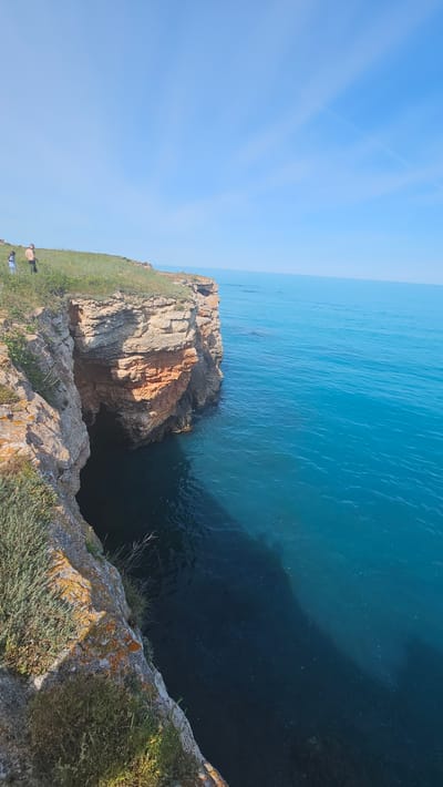The image depicts a coastal scene near Sveti Nikola, Bulgaria. A rocky cliff descends into the turquoise sea. The cliff is covered with green grass and some vegetation with orange lichen on the edge. A small rock formation with a red-orange hue stands out. There are two distant figures on the left of the cliff. The sky is blue with faint contrails.