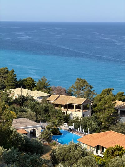 The image depicts an aerial view of a coastal landscape in the Municipal Unit of Sfakiotes, Greece. The Aegean Sea stretches along the left side of the frame with a gradient of blue shades near the horizon.  

On the right side, a series of buildings and trees line the coast. The architecture appears to be residential or resort-style with terracotta roofs. A clear, blue swimming pool is visible among the buildings, bordered by lounge chairs and umbrellas. Lush vegetation surrounds the structures.