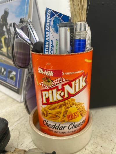 The image displays a close-up, eye-level shot of various items on a light-colored, textured surface, likely a desk. The primary focus is an orange cylindrical container of Pik-Nik Cheddar Cheese Shoestring Potatoes. The container is open at the top, revealing several items nestled inside, including what appears to be a small bunch of dry grass or straw, a silver cylindrical object with blue accents, and a metallic object. Partially obscuring the container is a pair of sunglasses with thick, clear frames and dark lenses. To the left of the sunglasses, a calendar with a motorcycle image is visible. Behind the Pik-Nik container, a blue and white card with text that includes "ICATION AND SERVICES" and "vega" is placed vertically. The overall lighting suggests an indoor setting, possibly an office or personal workspace. The background is dark and out of focus. The scene suggests a cluttered but organized desk setup. The location is identified as San Jose del Monte, Philippines.
