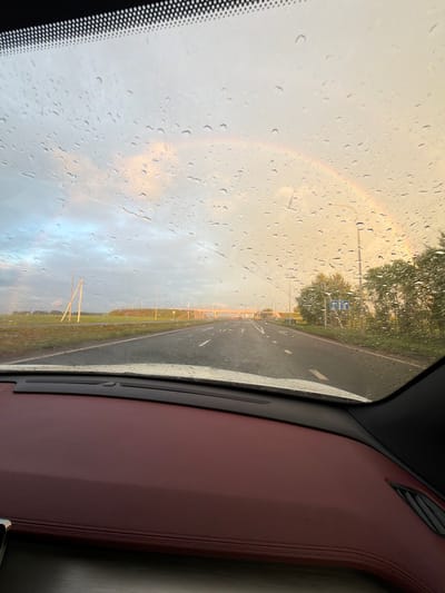 The image captures a view through a car windshield, displaying raindrops and a partial rainbow in a sky with soft, warm lighting. The road ahead appears wet, with white lane markings visible. On the sides of the road, there are fields, trees, and utility poles, along with a road sign indicating public transport facilities. The interior of the car shows a dark dashboard with burgundy leather accents. The scene suggests a drive through a rural or suburban area during or shortly after rainfall, under a dramatic sky. The location in Таймурзино, Russia, is mentioned, providing context to the geographical setting.
