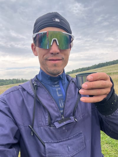 In an outdoor, rural setting resembling a meadow or field, a person is taking a selfie. They are wearing a black baseball cap and large, mirrored sunglasses with a white frame. The person is holding a small metal cup in their right hand and is wearing a dark blue hooded jacket over a blue athletic shirt. The background consists of a grassy field with scattered white flowers, a treeline in the distance, and a cloudy sky. There is no indication of the specific location within Auk-Bulyak, Russia.