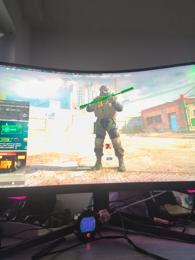 The image shows a monitor screen displaying a video game. The game appears to be a first-person shooter, featuring a character in military gear holding a green weapon. The character is standing in a desolate outdoor environment, with dilapidated buildings and a blurred background suggesting a post-apocalyptic or war-torn setting. On the left side of the screen, there are in-game menus and information panels, written in a Cyrillic alphabet. At the top of the screen, there are performance statistics showing CPU and GPU usage.
In the foreground, below the monitor, there is a desk cluttered with electronic devices and cables. A small round digital clock shows the date as 2026-05-19 and the time as 15:54. There's also a small figurine near the clock.
There is no indication of the location being Havixbeck, Germany, or any other specific location.