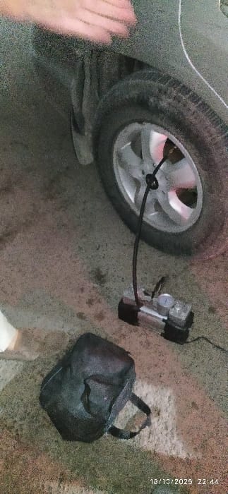 The image depicts a nighttime scene in Al-Kiswa, Syria, showing the lower rear section of a dark-colored vehicle with a dirty tire and silver-colored rim. A silver and black portable air compressor is positioned on the concrete or asphalt ground, connected by a black hose to the tire's valve stem, suggesting tire inflation is in progress. The compressor features a visible pressure gauge. To the left of the compressor, a dark-colored soft bag rests on the ground. A blurry, light-skinned hand is partially visible in the upper left quadrant, extended towards the car's fender. The ground is uneven and appears dirty with various stains. A timestamp in the lower right corner reads "18/10/2025 22:44".