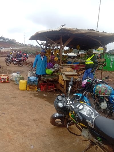 The image depicts an active outdoor market scene, likely a roadside fruit and vegetable stall, under an overcast sky. The ground is unpaved, appearing as dirt or reddish soil, possibly damp.
In the center of the image is a rustic stall constructed from wooden poles supporting a worn, light-colored canvas or tarp roof. Bunches of yellow bananas are clearly visible hanging from the stall's structure, indicating the primary goods sold. Beneath the roof, various other items, possibly produce, are arranged on makeshift tables or crates.
There are three main individuals associated with the stall. On the left, a man wearing a blue long-sleeved coat or overall and a cap stands facing the stall, holding a bright green bowl. His gaze is directed towards the goods or someone behind them. Under the stall, partially obscured by the hanging bananas, a woman in a yellow or orange top is visible, seemingly attending to the merchandise. To the right of the stall, a man in a dark jacket, blue pants, and a prominent green and yellow reflective vest stands, looking towards the stall or beyond it.
To the left of the stall, on the ground, are several containers including yellow jerry cans, blue plastic crates, and red plastic crates, alongside some large sacks or bags. Further into the background on the left, a group of people is gathered near several motorcycles, suggesting a common gathering point or a motorcycle taxi stand.
In the foreground, on the right side, two motorcycles are prominently displayed. The closest one is black and green, with "BOXER" visible on its fuel tank, which also features a white skull graphic and a "Monster Energy" logo. The handlebars and headlight are clearly visible. Behind it, a blue motorcycle has a colorful, possibly animated character graphic (resembling Ninja Turtles) on its tank.
In the background, on the far right, a green sign with white lettering is visible, bearing the text "Safaricom M-PESA," suggesting the location is likely in East Africa. The general background shows a hilly landscape with some buildings and what appears to be a cleared or constructed road area. The overall atmosphere is bustling but routine, characteristic of a local marketplace.