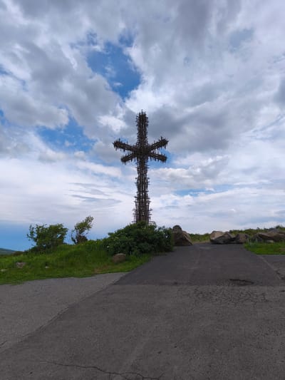 A tall, dark, cross-shaped structure, possibly under construction, dominates the center of the image. The cross is made of a dark metal framework, resembling scaffolding, and is silhouetted against a cloudy sky with patches of blue. The structure is situated on a grassy hilltop, with green bushes and trees visible around its base. In the foreground, a dark asphalt path, cracked and weathered in places, leads towards the cross. Several large, rough stones are scattered on the right side of the path. The scene appears to be an outdoor location, likely in a natural or elevated area, with a wide expanse of sky overhead. The overall impression is one of a monument in progress or an imposing landmark against a dynamic sky.