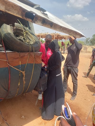 The image depicts an outdoor scene in Tataura, Nigeria, under a partly cloudy sky. The lower right foreground shows a person's hand holding a black wallet-style phone case containing a phone with a white charging cable.
The left side of the frame features the side of a heavily loaded vehicle, likely a truck or van. A spare tire is secured with yellow ropes atop large, dark bundled luggage, which is partially covered by a brown tarp also secured with ropes.
Three individuals are central to the composition. A male, wearing a red long-sleeved shirt with a white logo on the chest and white sneakers, is positioned near the vehicle, looking down and interacting with a female. This female, dressed in a black abaya with a black hijab and a silver-colored embellished band on her waistline, holds a black backpack or bag. A second male, wearing a black long-sleeved shirt with a white design, dark pants, and dark sneakers, stands slightly behind and to the right of the female. He holds a dark baseball cap on his head with his right hand.
In the mid-ground, behind the central figures, a rustic, open-sided shelter with a thatched roof supported by wooden poles is visible. Several other individuals are partially visible around and within this shelter and to the far right. The ground is reddish-brown earth. In the background, a light-colored wall or structure is present, and distant hills or elevated terrain are visible.